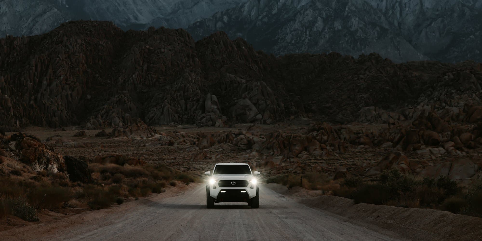 White Toyota Tacoma with its headlights on, driving down a desert road toward granite mountains at dusk