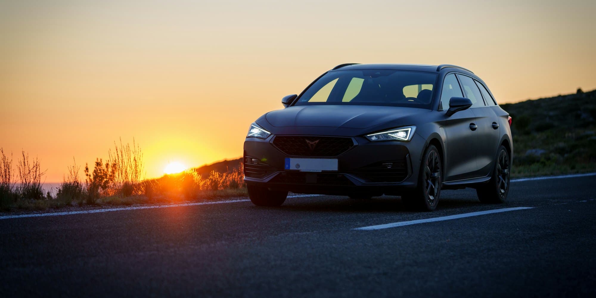Dark gray wagon parked on a coastal road as the sun sets behind it